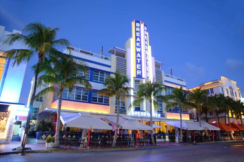 The Breakwater Hotel in South Beach at dusk, with its white and blue Art Deco facade and a prominent neon "BREAKWATER" sign on a vertical spire, with palm trees and a street-level restaurant.