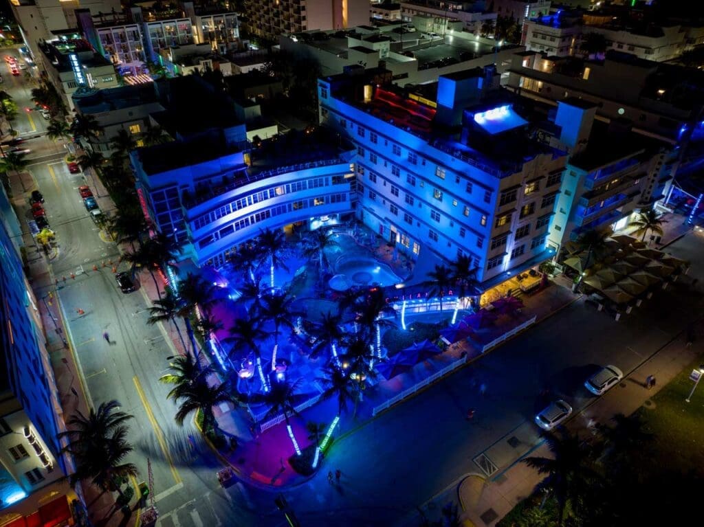 An aerial night view of The Clevelander Hotel in South Beach, with its distinctive art deco architecture, pool, and palm trees illuminated in vibrant neon blue and purple lights. The surrounding streets and other buildings are also visible.