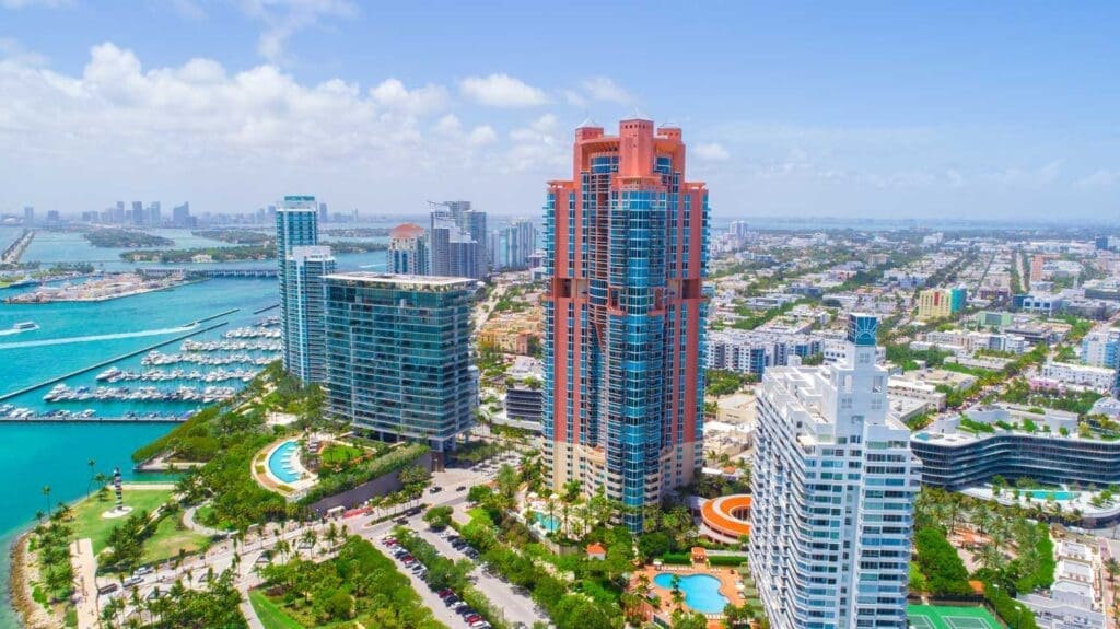 An expansive aerial view of the South of Fifth (SoFi) neighborhood in Miami Beach, with a prominent orange high-rise building at the center. The image showcases a marina, a golf course-like area, and numerous other buildings, with the Miami skyline visible across the turquoise bay in the distance.