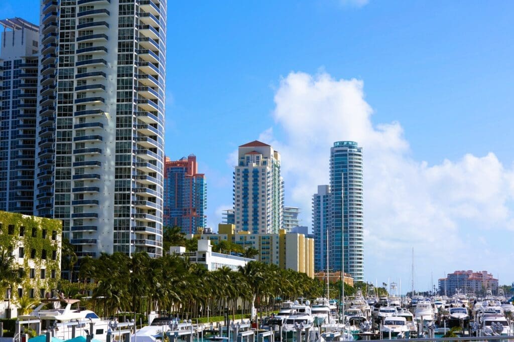A view of the Miami Beach Marina filled with boats and yachts, with the upscale buildings of South Beach in the background under a blue sky.