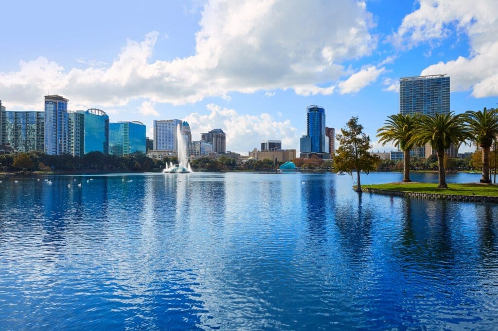 The Orlando skyline is seen across Lake Eola, with a fountain in the middle and a small island with palm trees on the right.