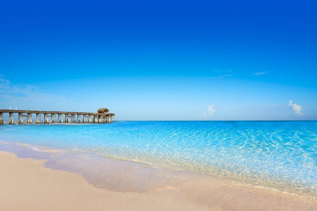 The Cocoa Beach Pier stretches over crystal-clear turquoise water, with a wide sandy beach in the foreground under a clear blue sky.