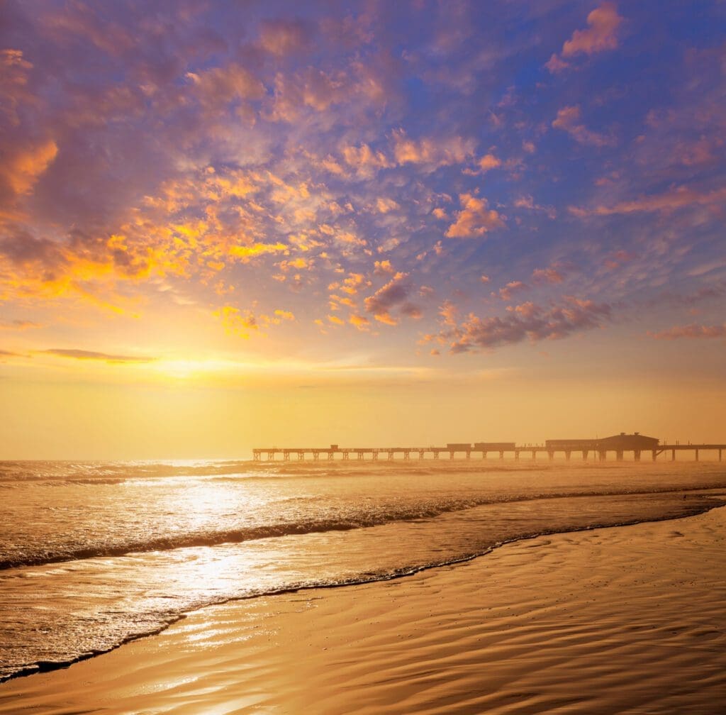 The pier at Daytona Beach is silhouetted against a vibrant orange and blue sunset sky, with the waves of the ocean rolling onto the sandy beach.