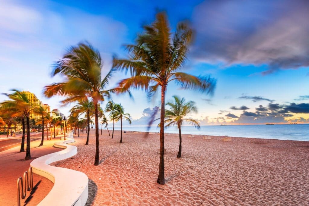A long exposure photo of Fort Lauderdale Beach at sunset, with blurred palm trees, a winding walkway, and the ocean in the background.