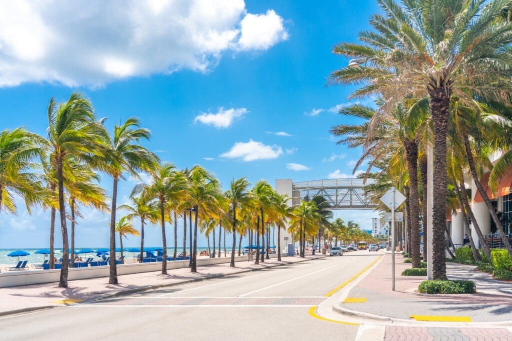 A street on the Fort Lauderdale Beach promenade lined with palm trees, with a beach and people under umbrellas on the left.