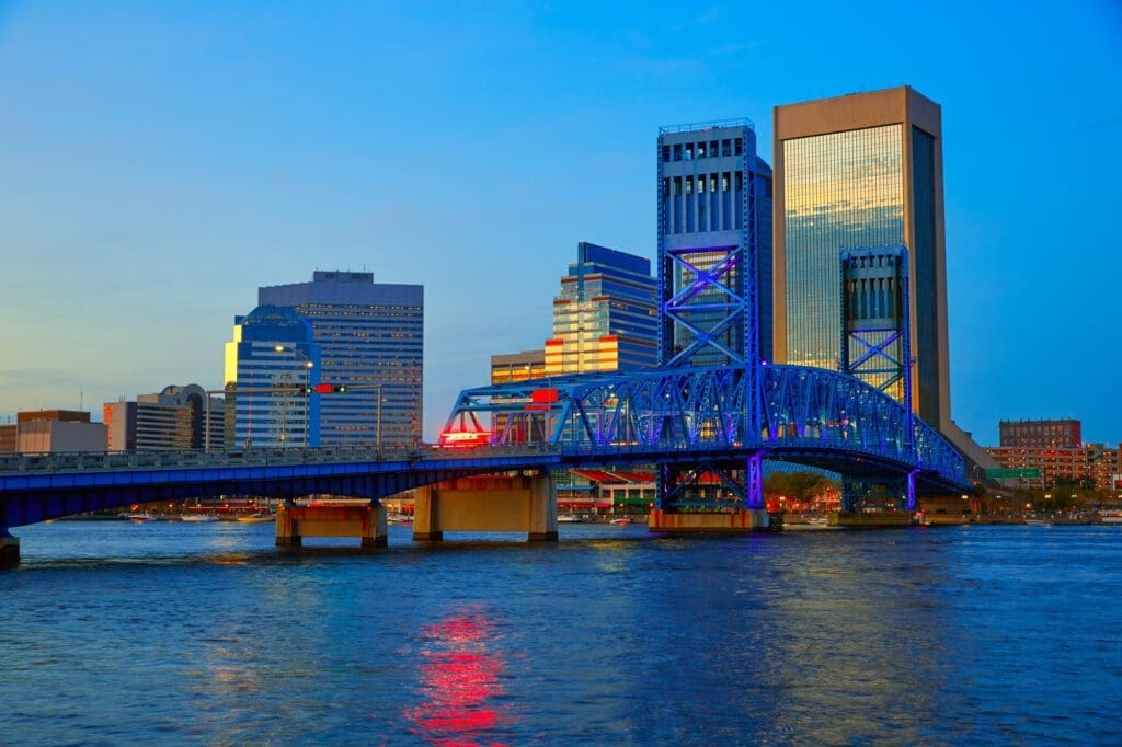 The Jacksonville skyline with a blue truss bridge over a river, with the buildings and bridge lights reflecting on the water in the evening.