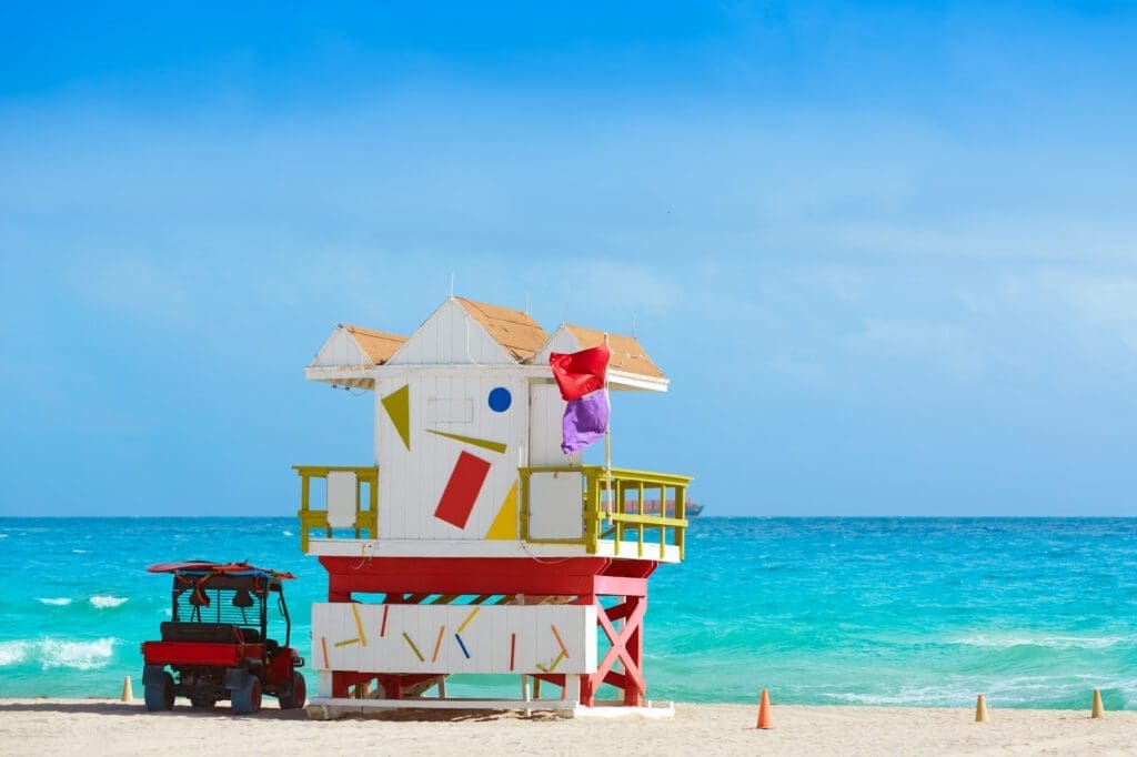 A colorful lifeguard tower with a red base and geometric shapes stands on the sand of South Beach, with a red ATV next to it and a turquoise ocean in the background.