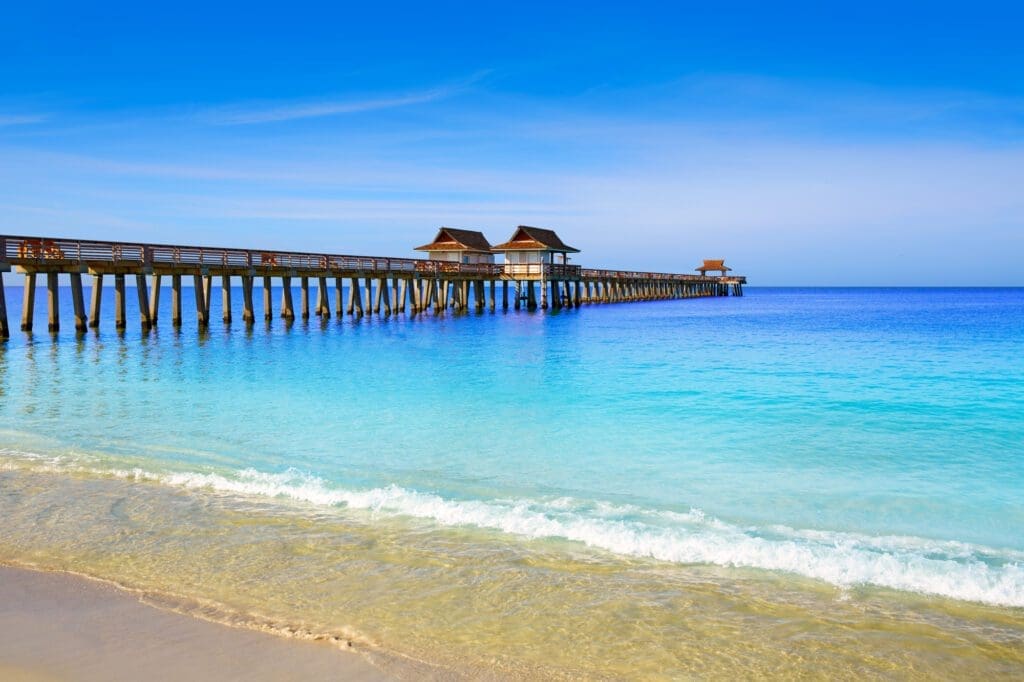 A view of the Naples Pier stretching over shimmering turquoise waters, with a wide sandy beach in the foreground under a blue sky.