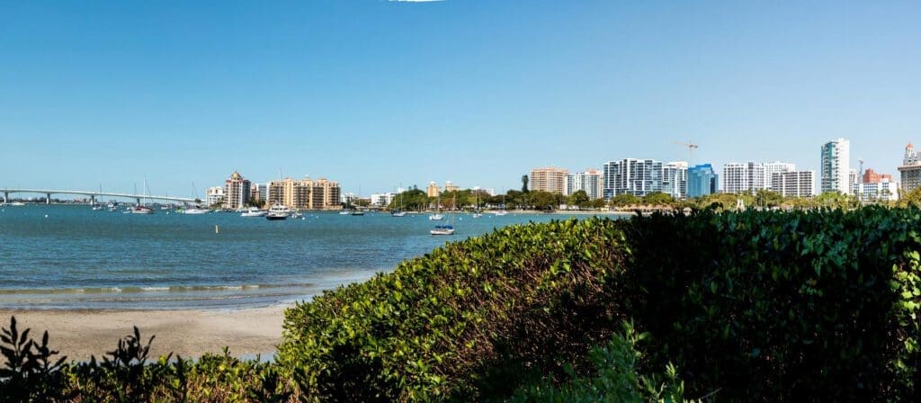 Sarasota Bay with a city skyline and the John Ringling Causeway Bridge, with boats and lush green vegetation in the foreground.