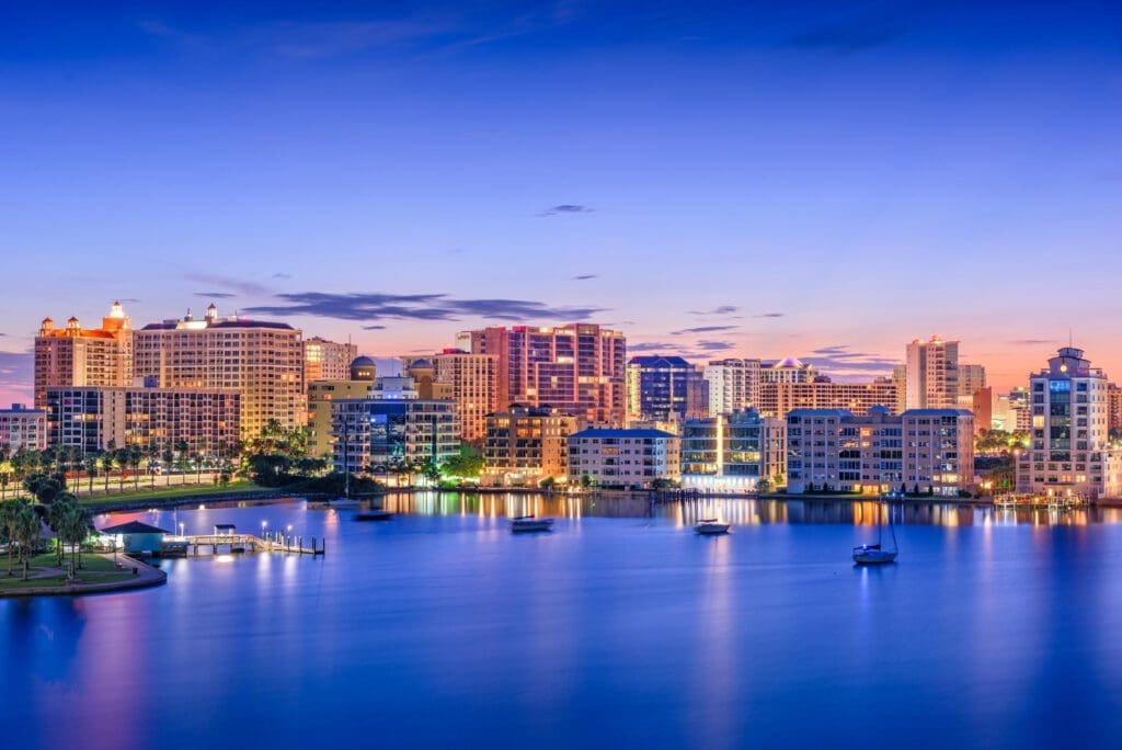 The Sarasota, Florida skyline with its lit buildings is seen at dusk from across a calm bay with boats.