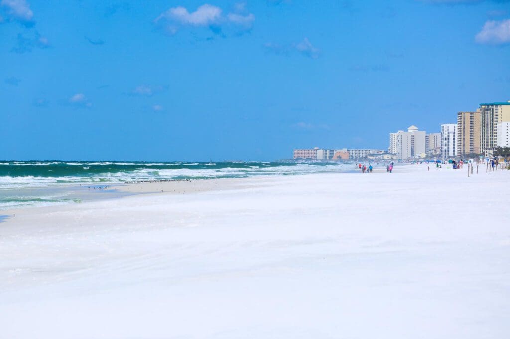 A wide, scenic view of the white sand beach and turquoise water in Destin, Florida, with high-rise buildings in the distance.