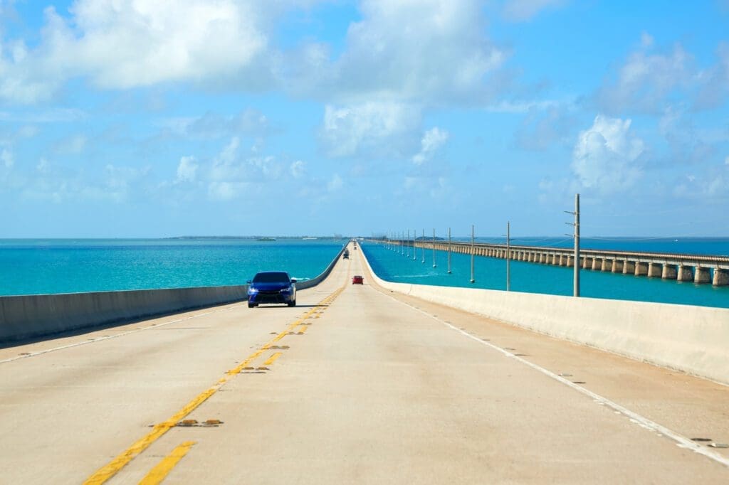 A scenic view of U.S. Highway 1 in the Florida Keys, a long bridge over a turquoise ocean, with a blue car driving in the foreground.