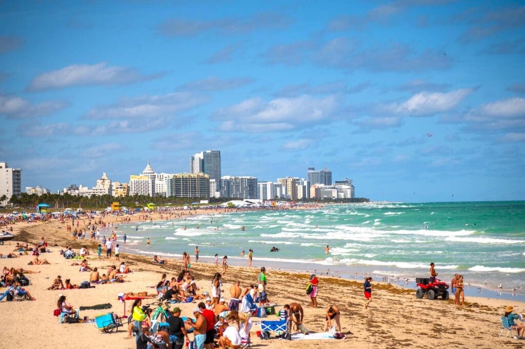 A wide, bustling view of South Pointe Beach in Miami Beach, with numerous people enjoying the sand and turquoise ocean waves, and the city skyline in the background.