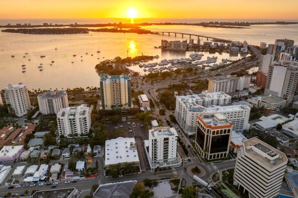 An aerial view of the Sarasota skyline and marina at sunset, with the sun setting over the bay and a bridge in the distance.