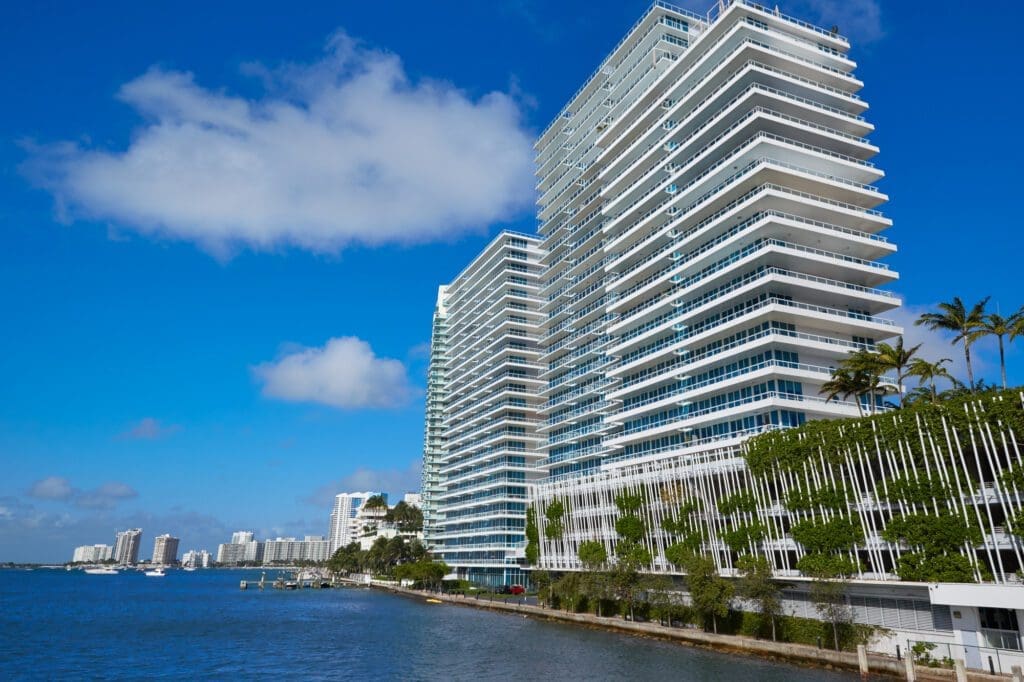 A view of modern high-rise buildings and palm trees in Miami Beach from across a bay under a blue sky.