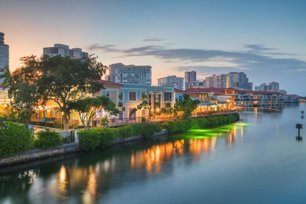 The waterfront skyline of Naples, Florida, with lit buildings and their reflections in a calm canal at dusk.