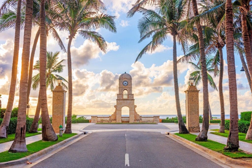 The Worth Avenue Clock Tower is seen at the end of a street lined with palm trees, with the ocean visible through its archway.