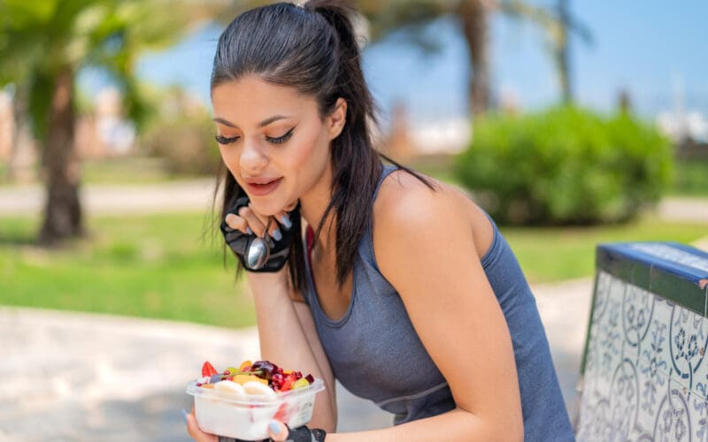 A young, fit woman is holding a bowl of fruit outdoors, preparing to enjoy a healthy snack after her workout