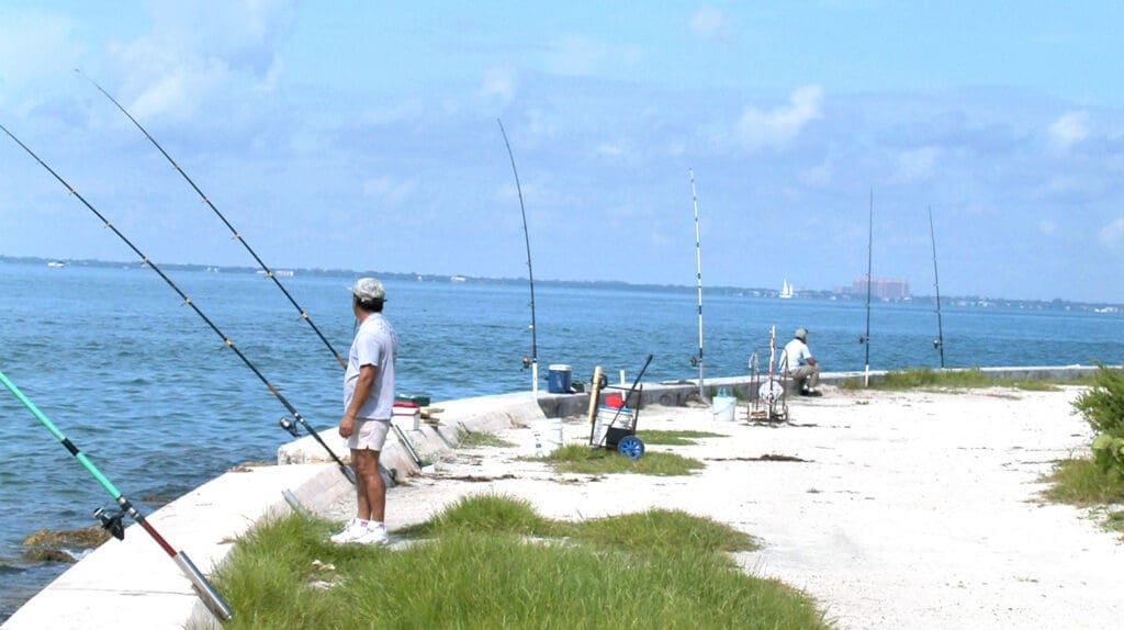 Two people are fishing on a concrete jetty at Bill Baggs Cape Florida State Park. One person in a white shirt stands with multiple fishing rods, while another is seated further down the jetty, with the bay and distant buildings under a partly cloudy sky.