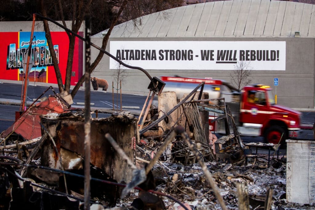 A fire truck passes by banner near structures damaged by Eaton Fire in Altadena