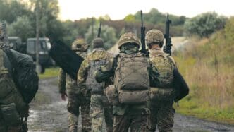 Military Personnel Walking on Rural Road. A group of military men returning after completing a task. Soldiers in military uniform with machine guns walk on a stony road with their backs to the cell.