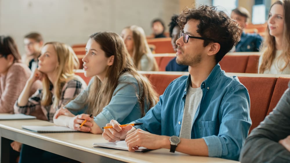 In the Classroom Multi Ethnic Students Listening to a Lecturer and Writing in Notebooks. Smart Young People Study at the College. By Shutterstock.com - Gorodenkoff