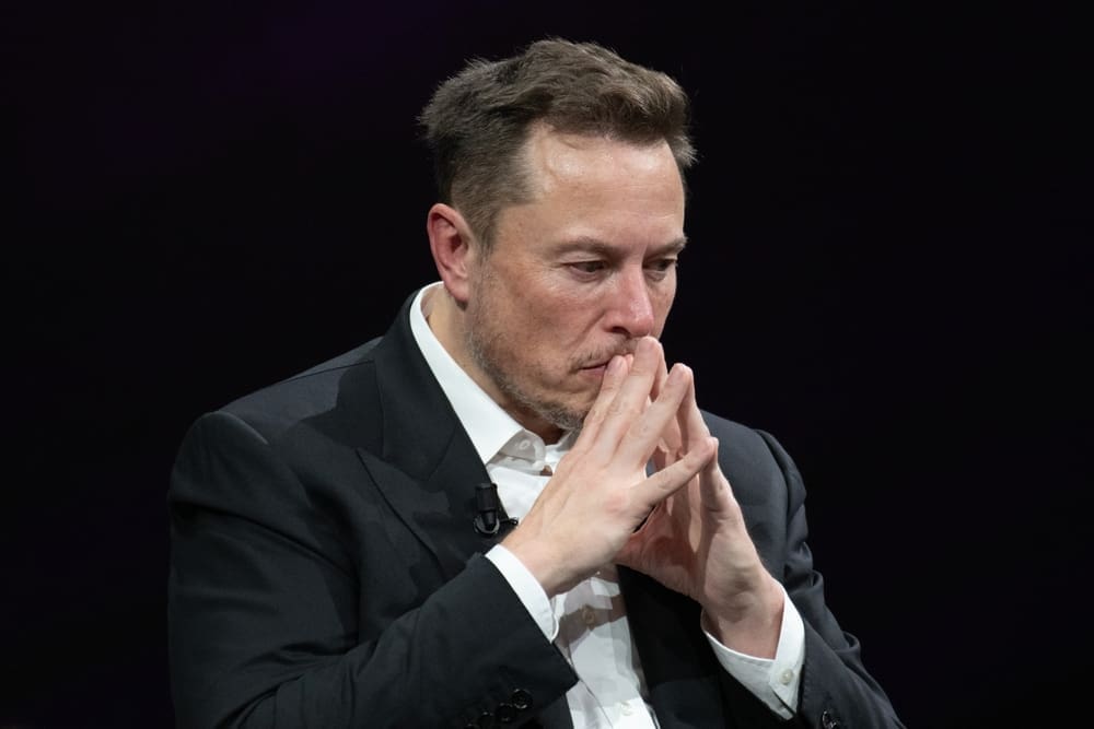 A close-up portrait of Elon Musk looking downwards with a pensive expression, his hands clasped together near his mouth, against a dark, plain background.