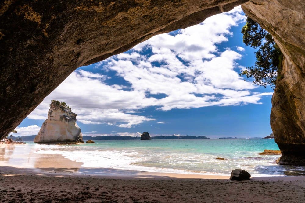 A stunning view through a large rock archway opening onto a sandy beach and turquoise ocean, with a distinctive sea stack and distant islands under a bright blue sky with white clouds at Cathedral Cove.