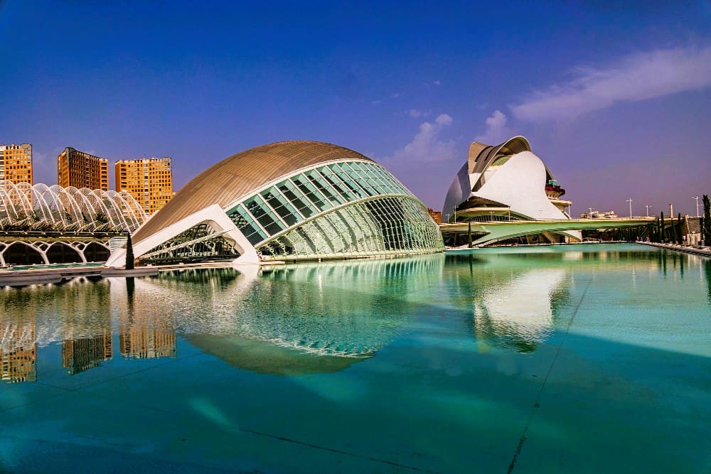The futuristic architecture of the City of Arts and Sciences in Valencia, Spain, including the Hemisfèric and Palau de les Arts Reina Sofía, is reflected in a large pool of turquoise water under a clear blue sky.