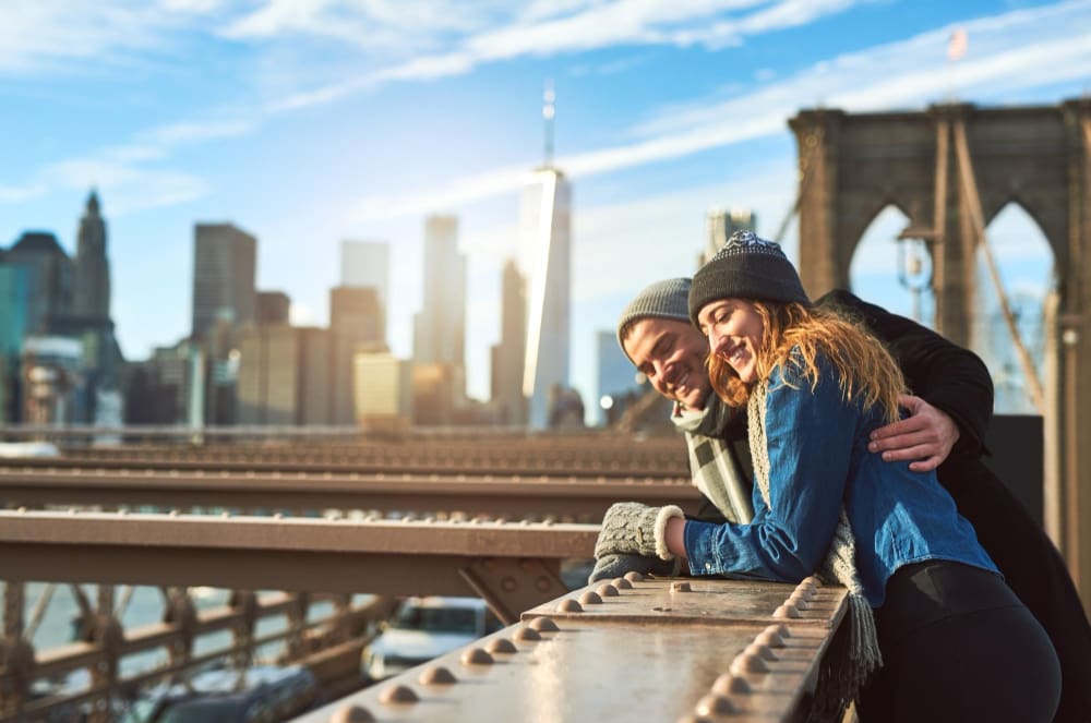 A happy couple, dressed in warm winter clothing, leans on the railing of the Brooklyn Bridge, looking at the New York City skyline with the One World Trade Center building prominent in the background.