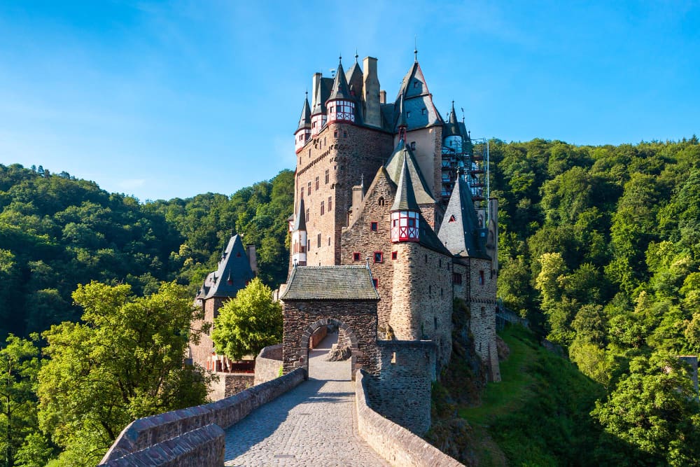 A magnificent view of Eltz Castle, a medieval fortress with multiple towers and red-and-white timber accents, accessed by a stone bridge, surrounded by lush green forests under a clear blue sky.