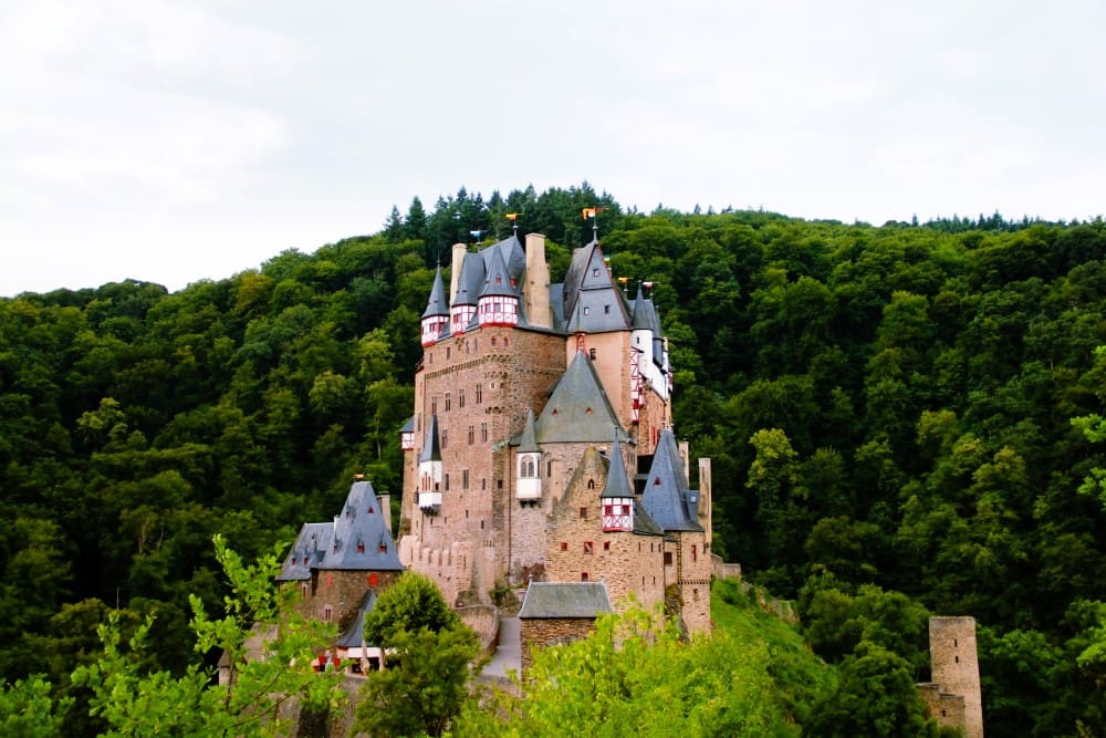 Eltz Castle, a medieval fortress with multiple towers and red-and-white accents, nestled amidst a dense green forest under a cloudy sky.