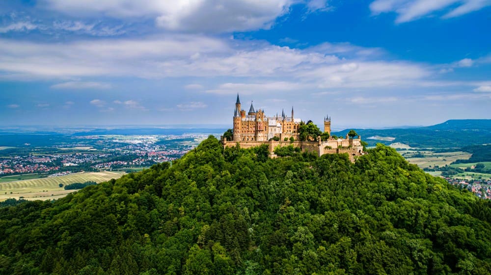Hohenzollern Castle, a majestic and imposing fortress with numerous towers, stands atop a verdant, tree-covered mountain, overlooking a vast landscape under a partly cloudy sky.