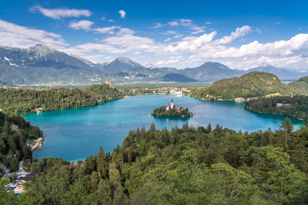 Panoramic view of Lake Bled in Slovenia, with its iconic island church and castle, surrounded by lush forests and mountains under a blue sky.