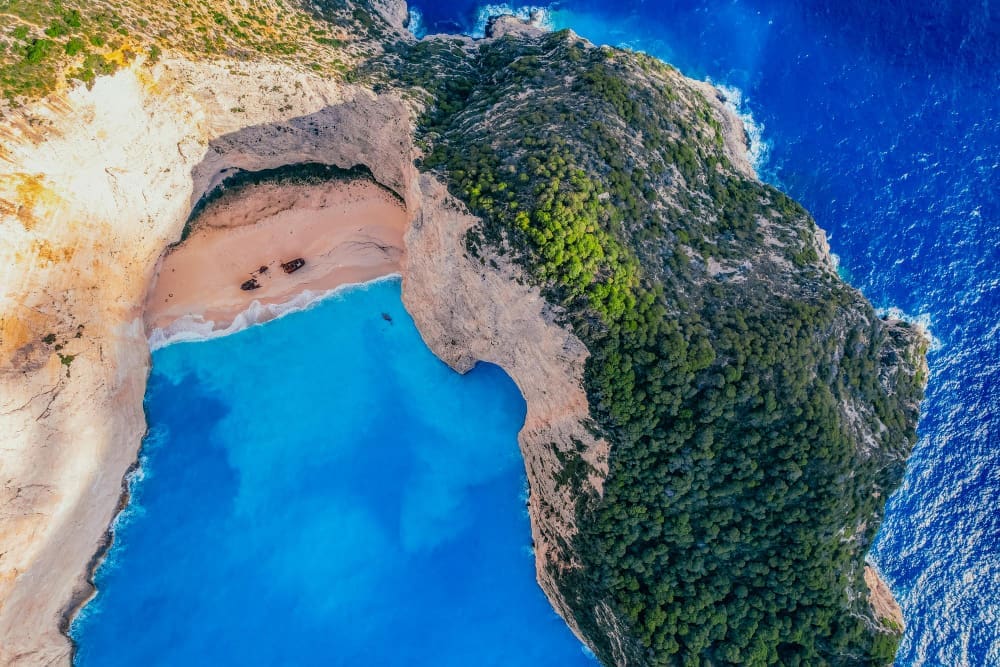 An aerial view looking down into Navagio Beach (Shipwreck Beach) in Zakynthos, Greece, showing its white sand, incredibly turquoise waters, towering white cliffs, and the famous shipwreck on the shore.