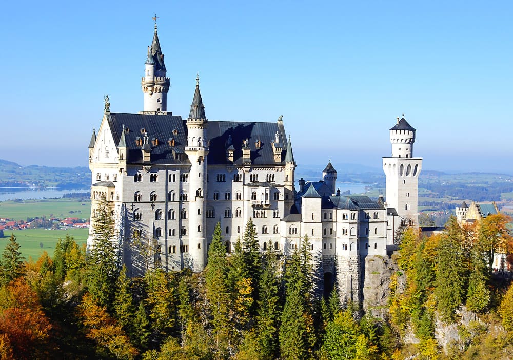 The fairytale Neuschwanstein Castle, a grand white stone structure with dark roofs and multiple turrets, perched atop a forested hill with vibrant autumn trees, under a clear blue sky.