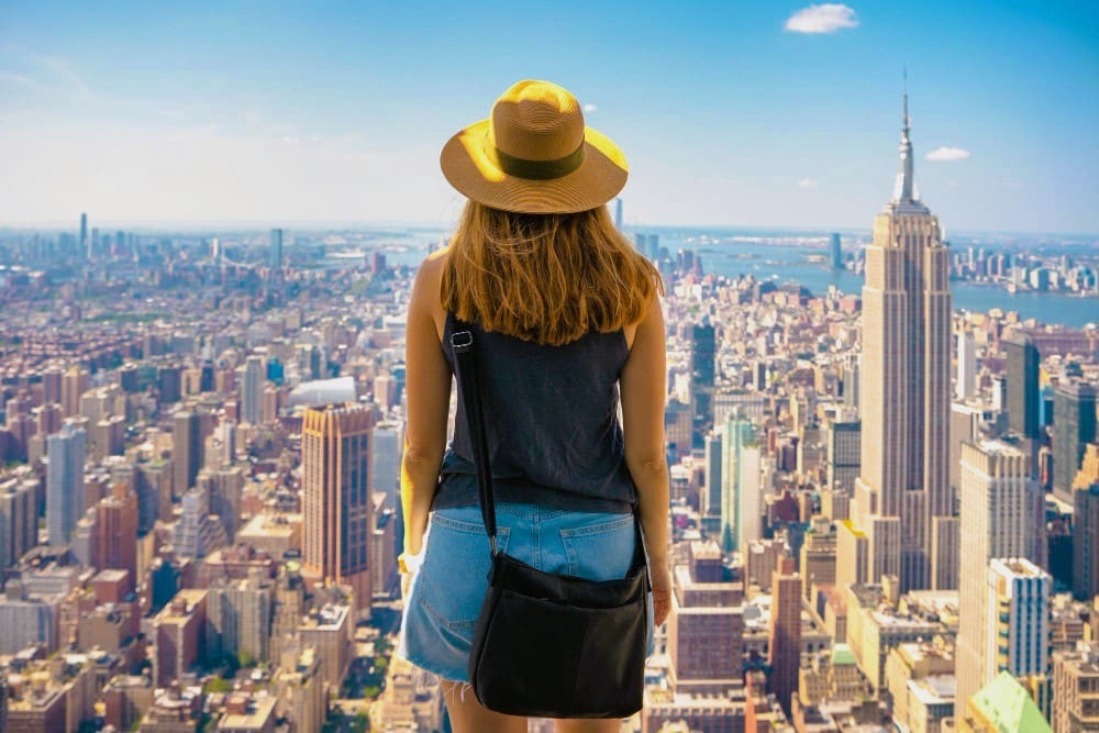 A woman in a straw hat and denim shorts, seen from behind, looks out over the expansive New York City skyline, with the Empire State Building prominent on the right, and the Statue of Liberty faintly visible in the distance.