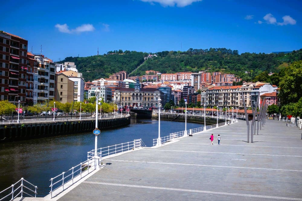 A wide view of the Nervión River in Bilbao, Spain, with a wide, empty promenade on the right and traditional buildings lining the riverbanks, backed by green hills under a bright blue sky.