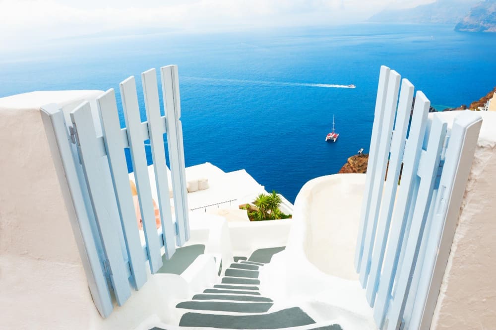 A classic view from Santorini, Greece, showing open light-blue wooden gates framing a whitewashed staircase leading down to the deep blue Aegean Sea, with a boat in the distance.