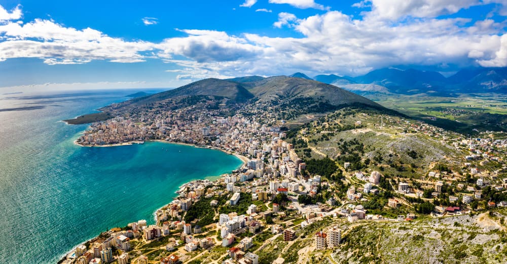 Aerial panoramic view of Sarandë, Albania, a coastal city nestled between mountains and the Ionian Sea, under a blue sky.