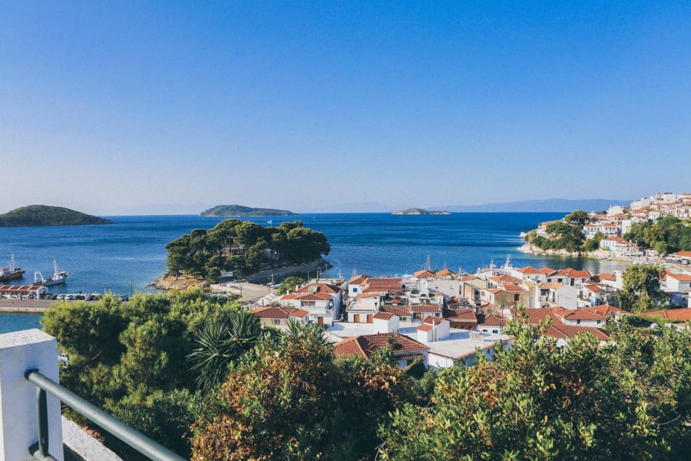An elevated view of Skiathos Town, Greece, showing white and brown buildings with red roofs clustered around a bay with small islands, surrounded by green trees and the clear blue Aegean Sea.