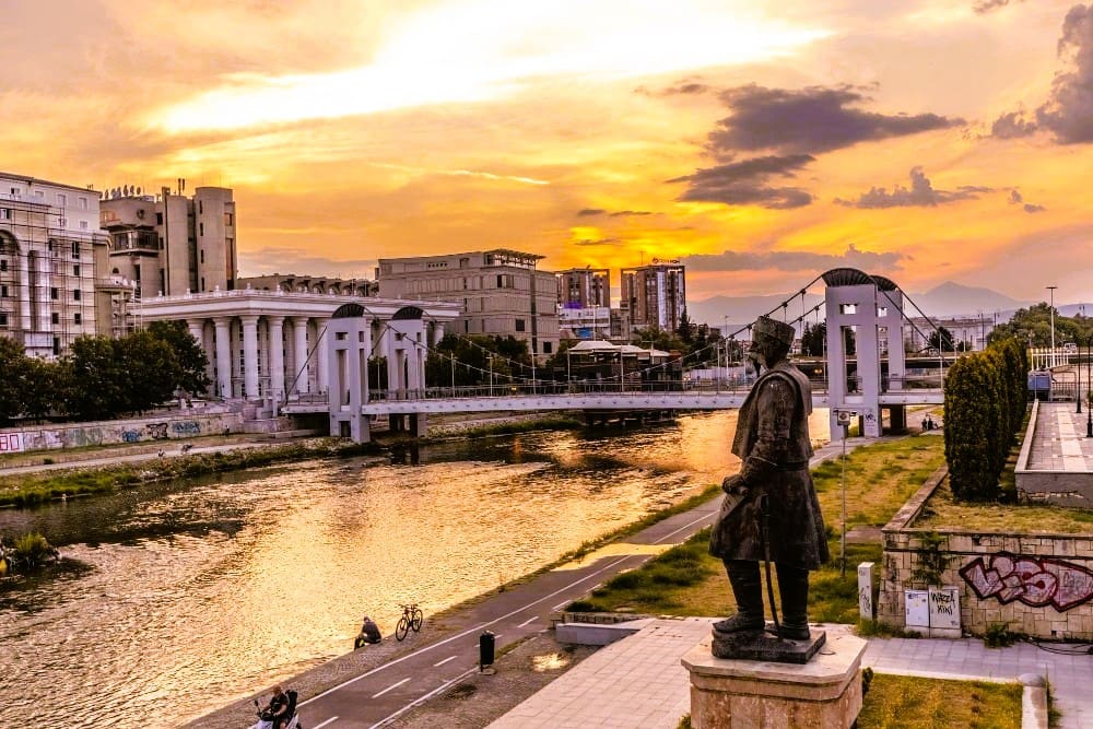 A statue overlooking the Vardar River in Skopje, North Macedonia, with the Art Bridge and city buildings at a vibrant sunset.