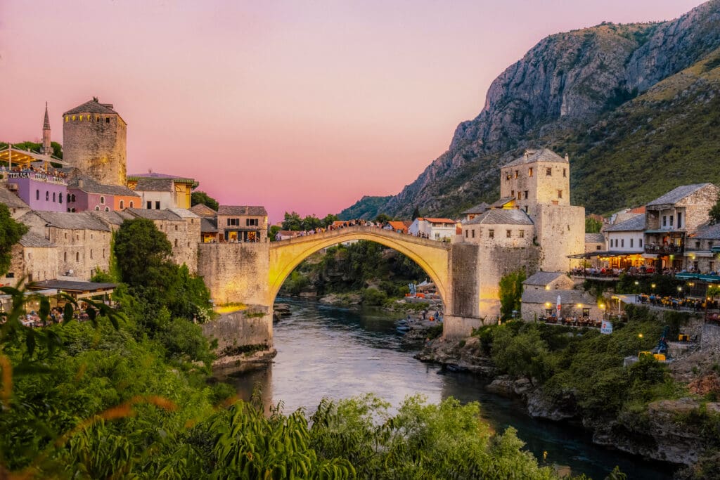 The illuminated Stari Most (Old Bridge) in Mostar, Bosnia and Herzegovina, arching over the Neretva River at dusk, surrounded by historic buildings and mountains.