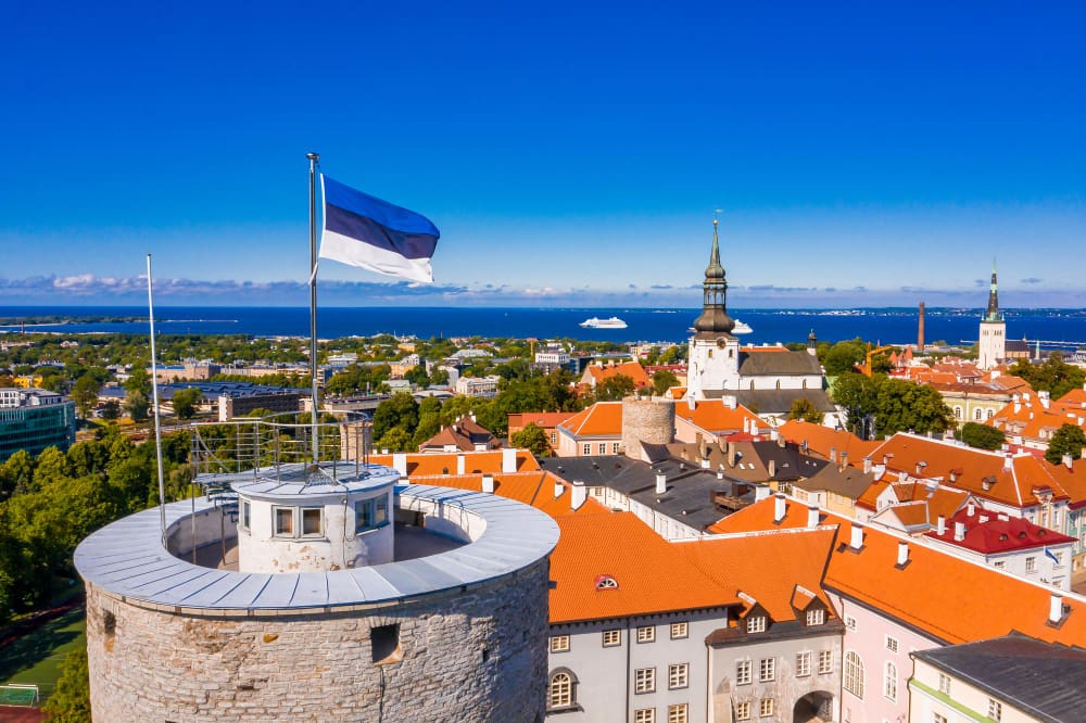 The Estonian flag flying atop Tall Hermann Tower in Tallinn Old Town, with the city and Baltic Sea in the background.