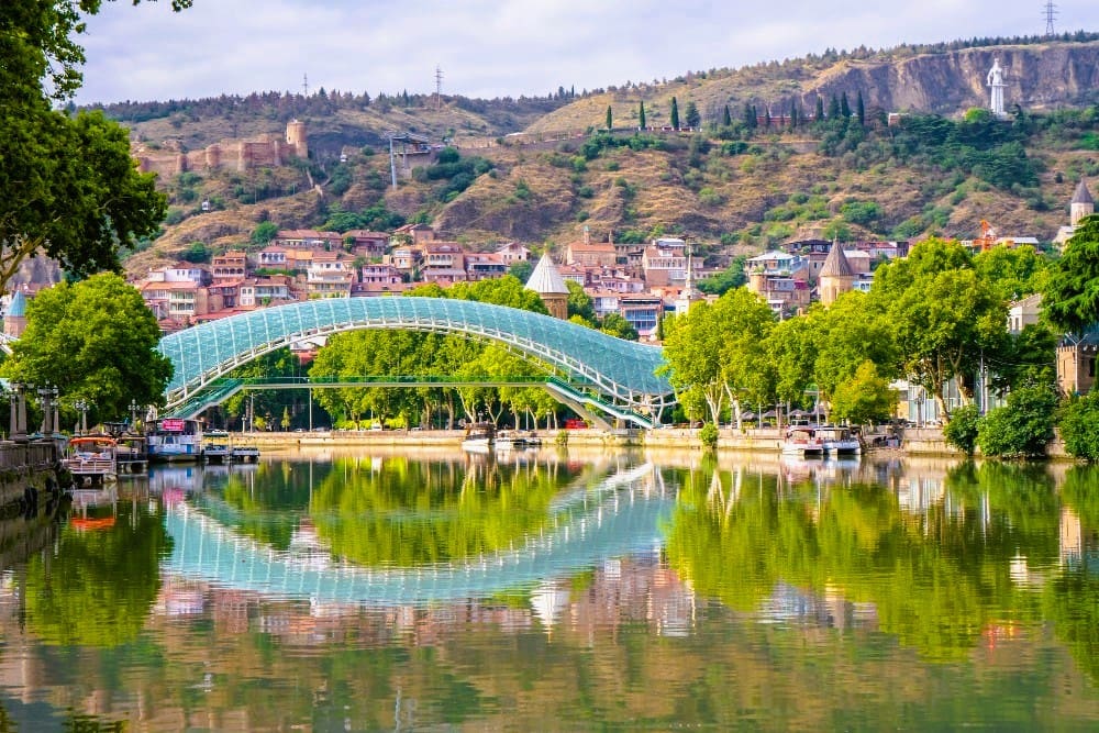 The modern, glass Peace Bridge spanning the Mtkvari River in Tbilisi, Georgia, with the old town and Narikala Fortress on the hillside.