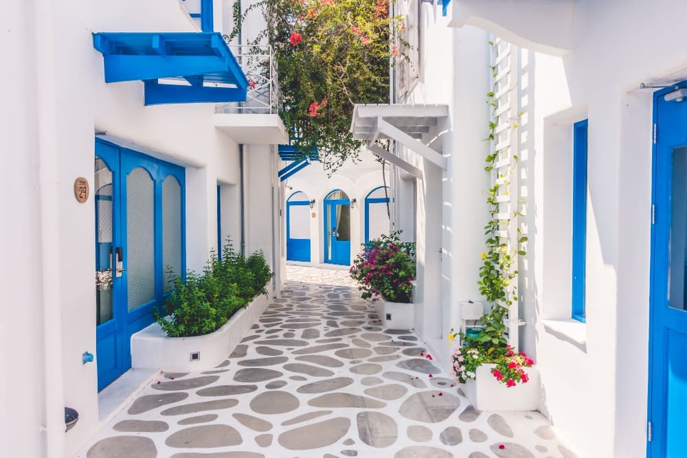 A narrow, whitewashed alleyway in Santorini, Greece, with bright blue doors and windows, potted plants, and irregular stone paving, characteristic of traditional Cycladic architecture.