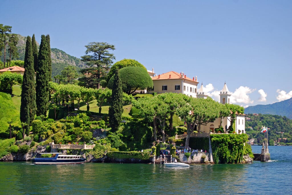 A panoramic view of Villa del Balbianello on Lake Como, Italy, featuring a grand white villa nestled in lush terraced gardens with cypress trees, overlooking the calm lake with docked boats.