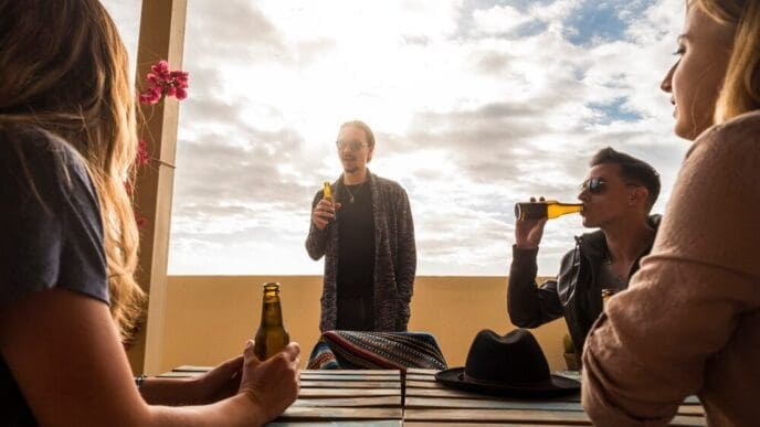 A group of young, white people socialize on a rooftop terrace, talking and drinking beer.
