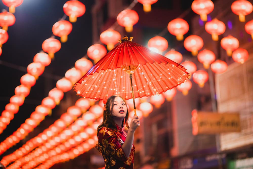 A woman in a red Chinese dress holds an illuminated red parasol under a string of glowing red lanterns at night.