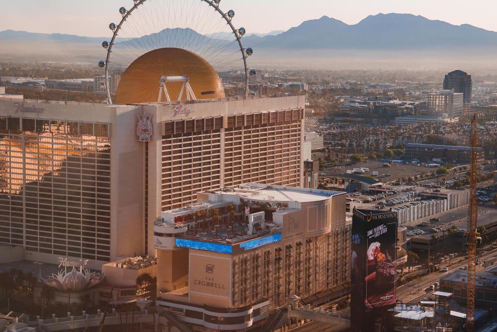 An aerial view of the Las Vegas Strip, showing the Flamingo and Cromwell hotels and the High Roller Ferris wheel with a mountain range in the background.